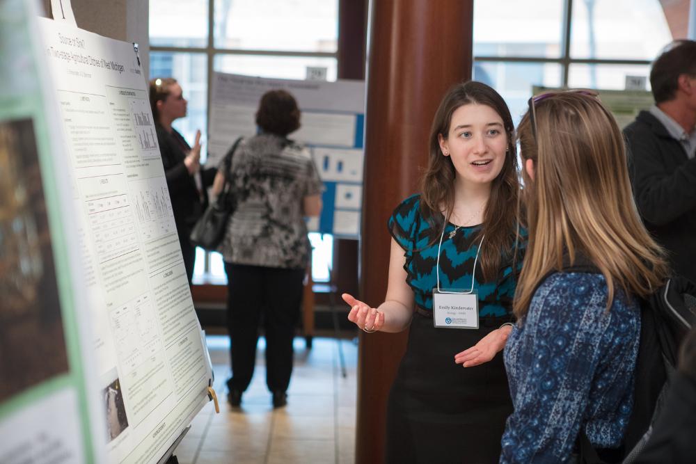 Student presenting their poster to an onlooker, gesturing with her hands